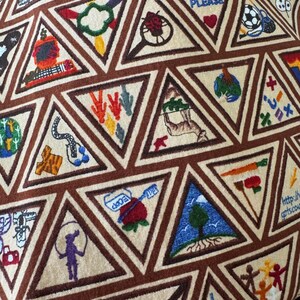 Puede incluir: Primer plano de una tela con un patr&oacute;n repetido de tri&aacute;ngulos bordados. Cada tri&aacute;ngulo contiene un dise&ntilde;o colorido diferente, incluyendo corazones, un bal&oacute;n de f&uacute;tbol y un &aacute;rbol. Los tri&aacute;ngulos est&aacute;n delineados en marr&oacute;n sobre un fondo beige.