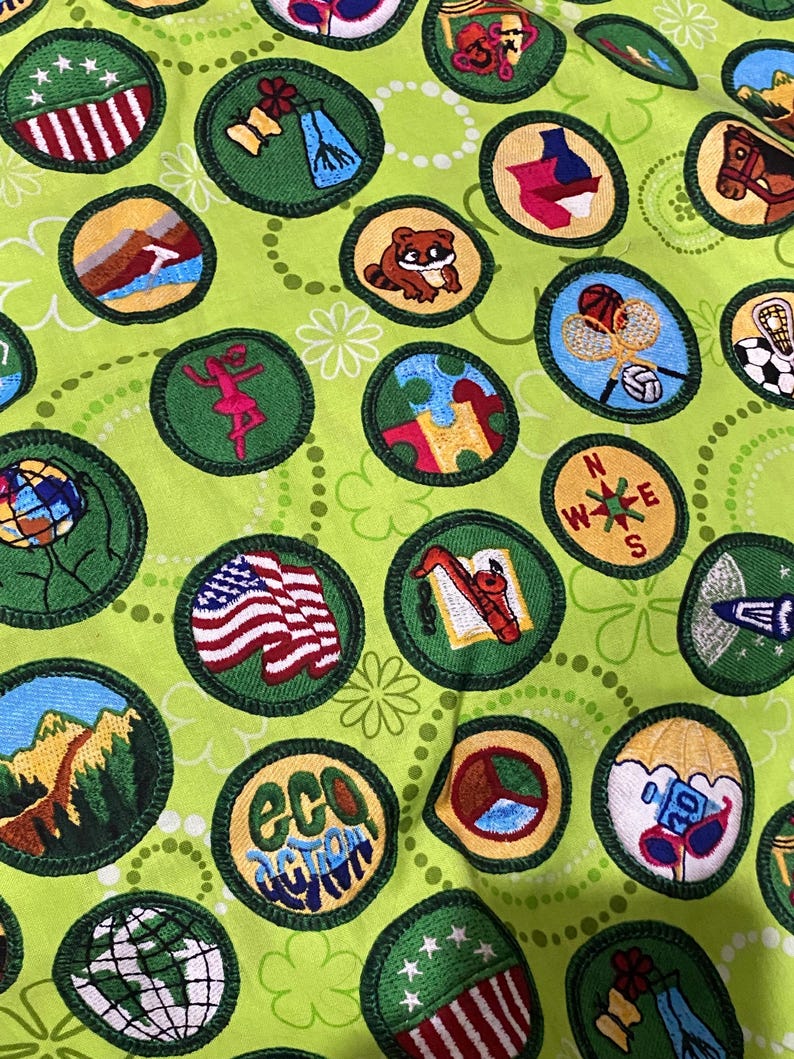 May include: Close-up of a fabric with various embroidered patches on a light green background. The patches feature images of a globe, a compass, a flag, and other designs. The patches are in a circular shape with a green border.