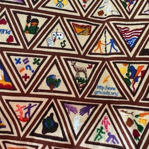 Puede incluir: Una tela beige con patrones triangulares marrones, cada uno con im&aacute;genes bordadas. Las im&aacute;genes incluyen un ciervo, manos, un globo y figuras tomadas de la mano. Tambi&eacute;n es visible la direcci&oacute;n del sitio web "http://www.gpts.scouts.org/".
