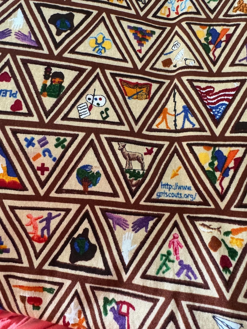 Puede incluir: Una tela beige con patrones triangulares marrones, cada uno con im&aacute;genes bordadas. Las im&aacute;genes incluyen un ciervo, manos, un globo y figuras tomadas de la mano. Tambi&eacute;n es visible la direcci&oacute;n del sitio web "http://www.gpts.scouts.org/".