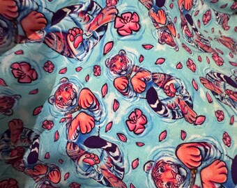 May include: A light blue fabric featuring a repeating pattern of tigers swimming in water, accented with scattered pink flower petals. The tigers are depicted with orange and black stripes, and each is shown floating with their paws visible.