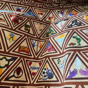 Puede incluir: Tela beige con un patr&oacute;n repetitivo de tri&aacute;ngulos marrones. Cada tri&aacute;ngulo contiene una imagen bordada diferente, como un bal&oacute;n de f&uacute;tbol, una bandera estadounidense y notas musicales. La palabra "PLEASE" es visible en un tri&aacute;ngulo.