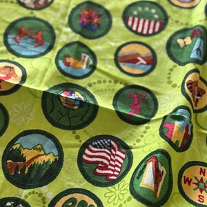 May include: A light green fabric with multiple embroidered patches. The patches are circular with various designs, including an American flag, mountains, and a compass. The fabric has a floral pattern.