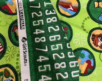 May include: A bright lime green fabric featuring a repeating pattern of circular embroidered-style patches with various designs. A white selvage edge displays the text Robert Kaufman, Girl Scouts, and Screen Print D# with a grid of numbers.