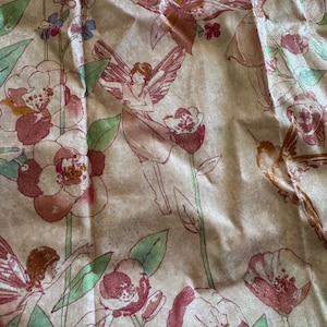 May include: A fabric with a repeating pattern of fairies, flowers, and hummingbirds in shades of pink, red, and green on a beige background. The design features detailed illustrations of floral elements and winged figures.