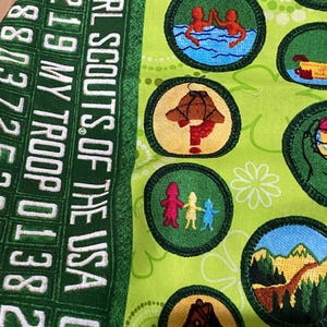 May include: Green fabric with embroidered Girl Scout badges. The badges feature various designs, including figures swimming, a mushroom, and a horse. The fabric also has the text "GIRL SCOUTS OF THE USA" and numbers.