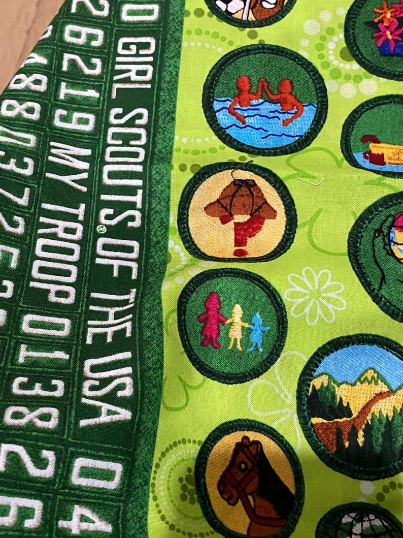 May include: Green fabric with embroidered Girl Scout badges. The badges feature various designs, including figures swimming, a mushroom, and a horse. The fabric also has the text "GIRL SCOUTS OF THE USA" and numbers.