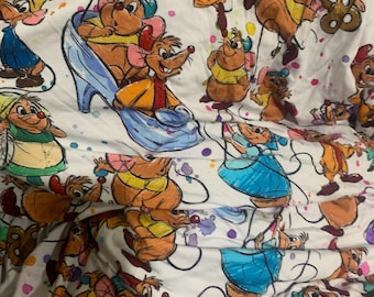 May include: A white fabric featuring a repeating pattern of cartoon mice in various poses and outfits, including yellow shirts and blue dresses. The design includes colorful paint splatter accents in shades of pink, purple, and green throughout the print.