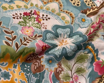 May include: Close-up of a floral fabric with a paisley design. The pattern features large teal flowers, pink and yellow blossoms, and green leaves on a light blue background. Brown and yellow accents add contrast to the colorful textile.
