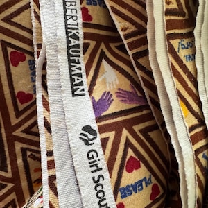 Puede incluir: Primer plano de tela con un patr&oacute;n geom&eacute;trico repetitivo en marr&oacute;n, beige y blanco. El patr&oacute;n incluye tri&aacute;ngulos, corazones y formas de manos. La tela tiene un borde blanco con el texto "ROBERT KAUFMAN" y "Girl Scouts&reg;".