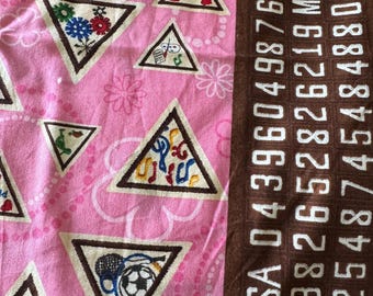 May include: Pink fabric with repeating triangular patches featuring various embroidered designs. The patches are outlined in brown and have a cream-coloured background. The right side of the fabric has vertical text and numbers in white on a brown background.