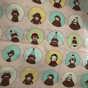 May include: Pink fabric with a repeating pattern of sock monkey faces inside of circles. The sock monkeys have red mouths and are wearing hats, party hats, and bow ties. The circles are in shades of yellow and blue.