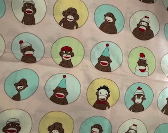 May include: Pink fabric featuring a repeating pattern of sock monkey faces within circles. The sock monkeys have red mouths and wear hats, party hats, and bow ties. The circles are in shades of yellow and blue.