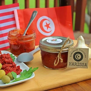 May include: Two glass jars filled with red harissa paste, one with a label that reads "Homemade Tunisian Harissa". The jars are on a wooden surface with a white plate of olives and red pepper flakes, and two flags, one American and one Tunisian.