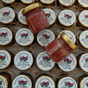 May include: A collection of small glass jars filled with red harissa paste, each with a gold lid. The jars are arranged in a box, surrounded by lids with the text "Extreme Harissa 2025" and a chili pepper graphic. The jars are tied with twine.