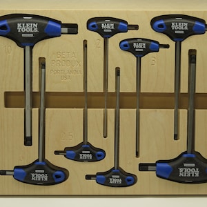 Op de afbeelding: Een set van acht Klein Tools T-greep inbussleutels met blauwe en zwarte handgrepen, georganiseerd in een lichtgekleurd houten opbergplateau. Het plateau bevat gegraveerde numerieke labels en de tekst BETA PRODUX PORTLANDIA USA.