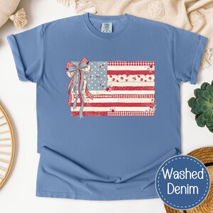 May include: A light blue t-shirt with a patriotic American flag design. The flag design includes a bow and ribbons, with red, white, and blue patterns. The shirt is made of washed denim. The t-shirt is a comfortable, casual wear item.