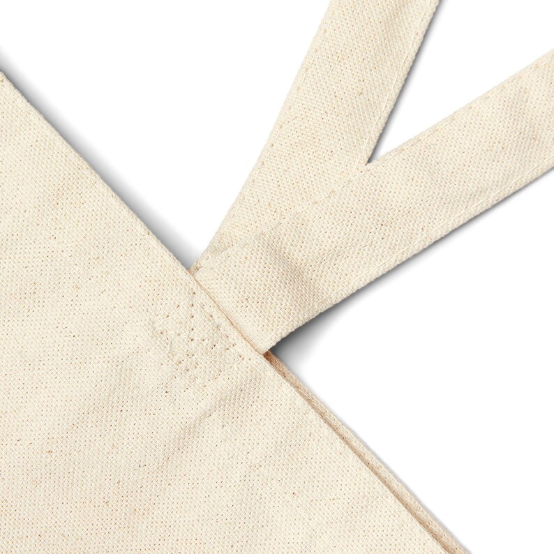 May include: Close-up of a natural canvas tote bag. The bag features a sturdy, cream-colored fabric with a visible weave. The straps are sewn securely to the bag, creating a durable design. The neutral color makes it versatile for various uses.