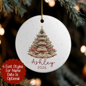 Puede incluir: Adorno navideño redondo de cerámica blanca con un diseño de árbol de Navidad hecho de libros. El adorno tiene el nombre "Ashley" y el año "2026" en escritura roja. El adorno cuelga de una cuerda dorada.