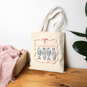 May include: Cream-colored tote bag with a red and blue illustration. The design features the words "La Dolce Vita" and "Sardinia" with four blue fish. The bag has two cream-colored handles.