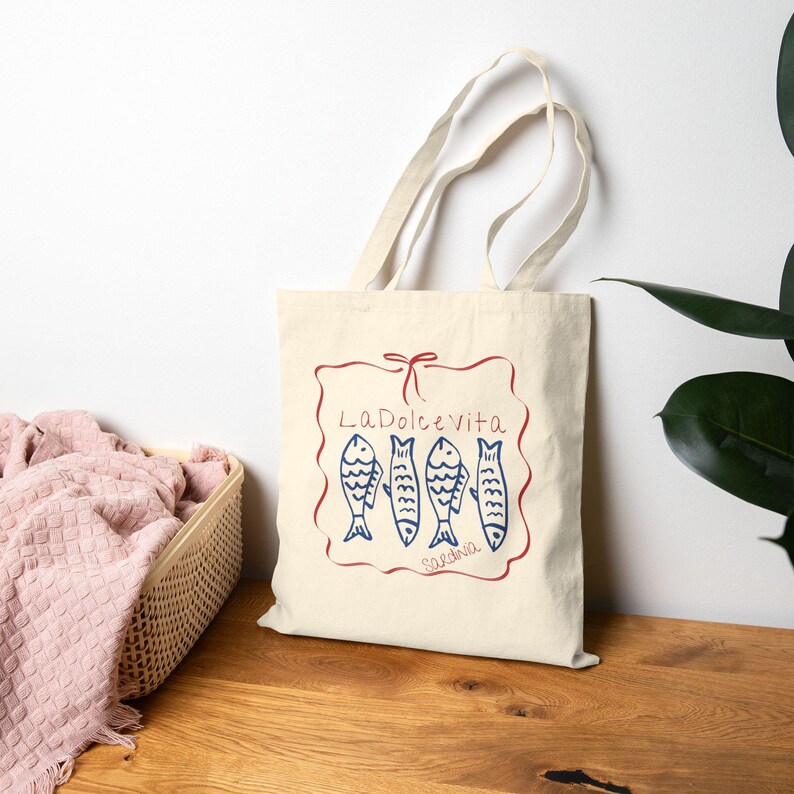 May include: Cream-colored tote bag with a red and blue illustration. The design features the words "La Dolce Vita" and "Sardinia" with four blue fish. The bag has two cream-colored handles.