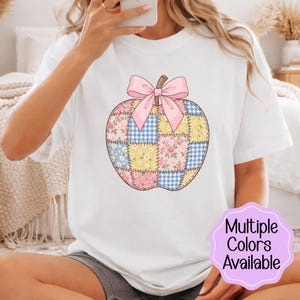 Peut inclure: Un t-shirt blanc à manches courtes avec un motif central représentant une pomme en patchwork. Le design de la pomme combine des carreaux vichy bleus, des fleurs jaunes et des roses roses, surmonté d'un nœud rose clair. Un badge violet indique Multiple Colors Available.