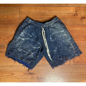 May include: Navy blue shorts with a distressed, frayed hem and white paint splatter details. The shorts feature a drawstring waist and a blue lining. The style is casual and artistic. The shorts are approximately 40 cm in length.