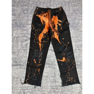 Könnte beinhalten: Schwarze Jeans mit orangefarbenen Farbspritzern und Distressed-Details. Die Jeans haben ein einzigartiges Design mit orangefarbener Farbe auf der Rückseite und Spritzern auf der Vorderseite. Die Jeans haben zerrissene Knie und einen rohen Saum.