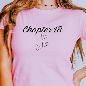 Peut inclure: Un t-shirt rose clair avec le texte "Chapter 18" en écriture noire, accompagné de deux petits cœurs. Le t-shirt est porté avec un jean bleu clair.