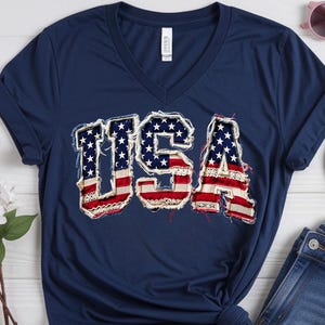 Könnte beinhalten: Ein marineblaues V-Ausschnitt-T-Shirt mit dem Text USA in einem abgenutzten amerikanischen Flaggenmuster mit ausgefransten Kanten. Das Shirt wird zusammen mit blauen Jeansshorts, blauen Flip-Flops und einer rosa Sonnenbrille gezeigt. Ein Bella Canvas Etikett ist sichtbar.