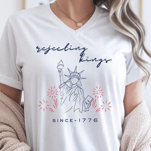 May include: A white V-neck t-shirt featuring a dark blue line art illustration of the Statue of Liberty, red firework accents, and the text "rejecting kings" in dark blue script above the statue and "SINCE-1776" in dark blue text below.