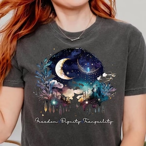 Può includere: T-shirt grigio scuro con un disegno ad acquerello di una luna crescente, stelle e un paesaggio celeste. La maglietta ha le parole "Freedom Dignity Tranquility" stampate in basso. La maglietta è realizzata in materiale morbido.