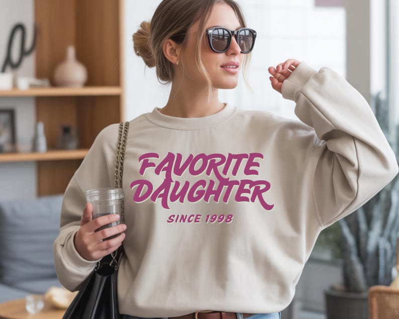 Op de afbeelding: Een beige sweatshirt met de tekst "Favorite Daughter" in een roze, retro lettertype. De tekst "Since 1998" staat eronder. De sweatshirt heeft een casual stijl.