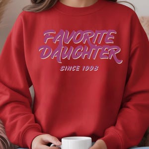 Op de afbeelding: Een rode sweatshirt met de tekst "FAVORITE DAUGHTER" in paarse en witte letters, met daaronder "SINCE 1993". De sweatshirt heeft een effen rode kleur.