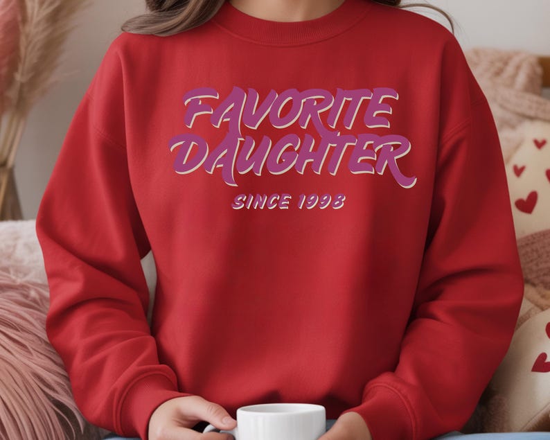Op de afbeelding: Een rode sweatshirt met de tekst "FAVORITE DAUGHTER" in paarse en witte letters, met daaronder "SINCE 1993". De sweatshirt heeft een effen rode kleur.