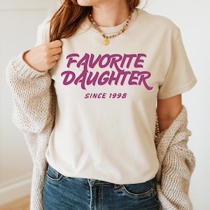Op de afbeelding: Een cr&egrave;mekleurig T-shirt met de tekst "FAVORITE DAUGHTER SINCE 1998" in een opvallend paars lettertype. Het shirt heeft een casual stijl, perfect voor dagelijks gebruik. De tekst is gecentreerd op de voorkant van het shirt.