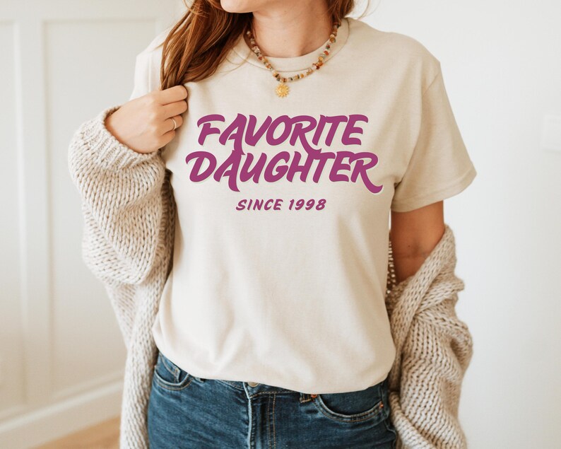 Op de afbeelding: Een cr&egrave;mekleurig T-shirt met de tekst "FAVORITE DAUGHTER SINCE 1998" in een opvallend paars lettertype. Het shirt heeft een casual stijl, perfect voor dagelijks gebruik. De tekst is gecentreerd op de voorkant van het shirt.