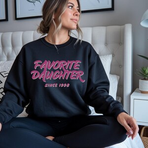 Op de afbeelding: Zwart sweatshirt met de tekst "FAVORITE DAUGHTER" in roze en wit, met "SINCE 1998" eronder. De sweatshirt heeft een ronde hals.