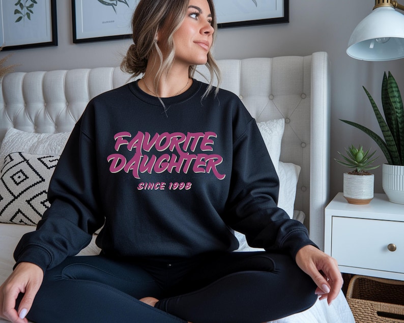 Op de afbeelding: Zwart sweatshirt met de tekst "FAVORITE DAUGHTER" in roze en wit, met "SINCE 1998" eronder. De sweatshirt heeft een ronde hals.