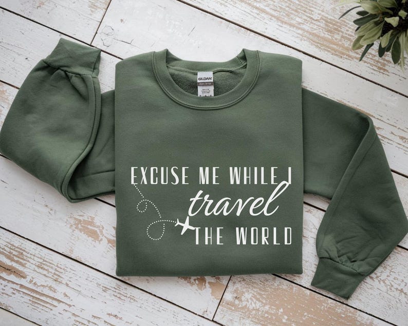 May include: Olive green sweatshirt with the text "EXCUSE ME WHILE I travel THE WORLD" in white. The design includes a dotted line and airplane graphic. The sweatshirt has a crew neck and long sleeves.