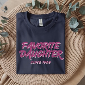 Op de afbeelding: Marineblauwe t-shirt met de tekst "Favorite Daughter" in roze en wit, met "Since 1998" eronder. De t-shirt ligt op een ronde, geweven ondergrond met decoratieve bladeren.