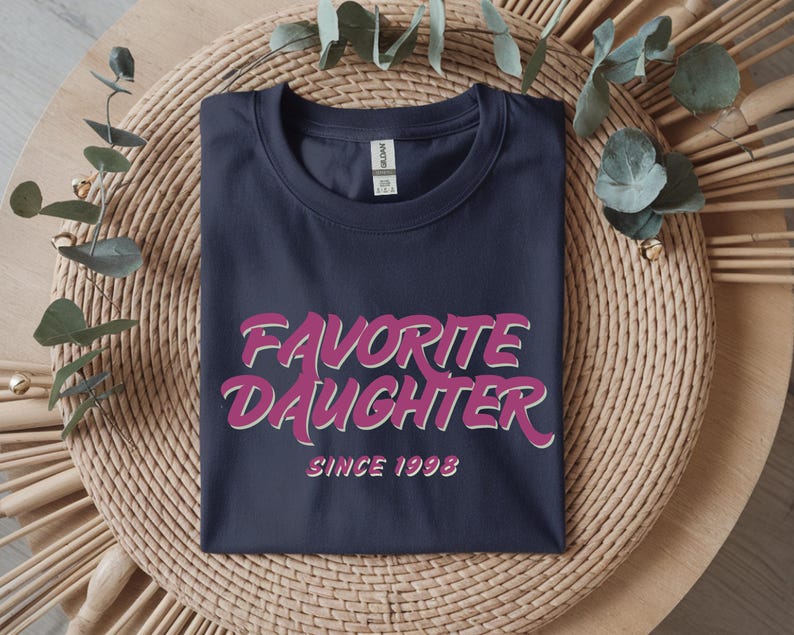 Op de afbeelding: Marineblauwe t-shirt met de tekst "Favorite Daughter" in roze en wit, met "Since 1998" eronder. De t-shirt ligt op een ronde, geweven ondergrond met decoratieve bladeren.