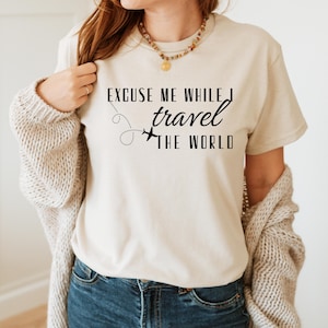 May include: Beige t-shirt with the text "EXCUSE ME WHILE I travel THE WORLD" in black. The design includes a dotted line and airplane graphic. The person is wearing blue jeans and a beige cardigan.