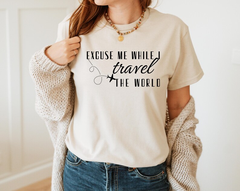 May include: Beige t-shirt with the text "EXCUSE ME WHILE I travel THE WORLD" in black. The design includes a dotted line and airplane graphic. The person is wearing blue jeans and a beige cardigan.