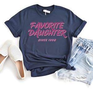 Op de afbeelding: Marineblauw T-shirt met de tekst "FAVORITE DAUGHTER SINCE 1998" in roze en witte letters. Het shirt wordt gecombineerd met een lichtblauwe spijkerbroek en witte instappers.