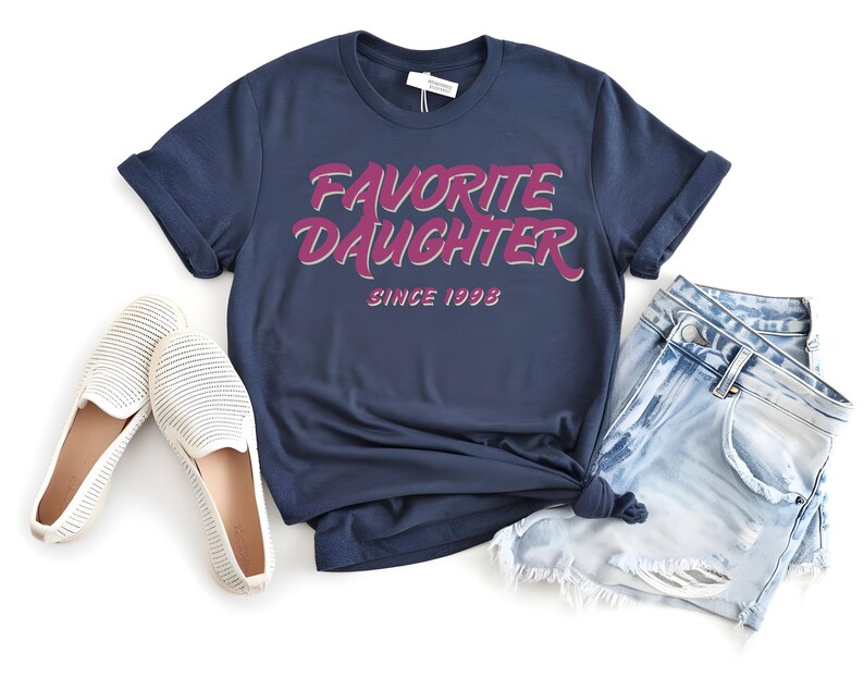 Op de afbeelding: Marineblauw T-shirt met de tekst "FAVORITE DAUGHTER SINCE 1998" in roze en witte letters. Het shirt wordt gecombineerd met een lichtblauwe spijkerbroek en witte instappers.