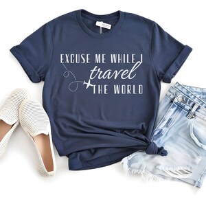 May include: Navy blue t-shirt with the text "Excuse Me While I Travel The World" in white. The shirt is paired with light wash denim shorts and white slip-on shoes. A travel-themed outfit.