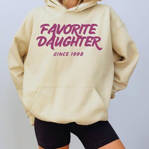 Op de afbeelding: Een beige hoodie met een voorzak en een capuchon met trekkoord. De hoodie heeft de tekst "FAVORITE DAUGHTER" in een cursief lettertype, met "SINCE 1998" eronder, allemaal in een dieproze kleur.
