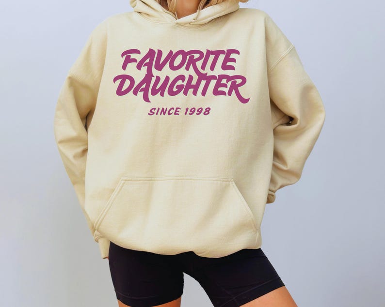 Op de afbeelding: Een beige hoodie met een voorzak en een capuchon met trekkoord. De hoodie heeft de tekst "FAVORITE DAUGHTER" in een cursief lettertype, met "SINCE 1998" eronder, allemaal in een dieproze kleur.