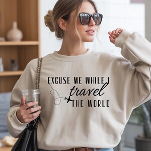 May include: A cream-colored sweatshirt with the text "EXCUSE ME WHILE I travel THE WORLD" in black. The design includes a dotted line and airplane graphic. The sweatshirt is a casual travel-themed apparel item.